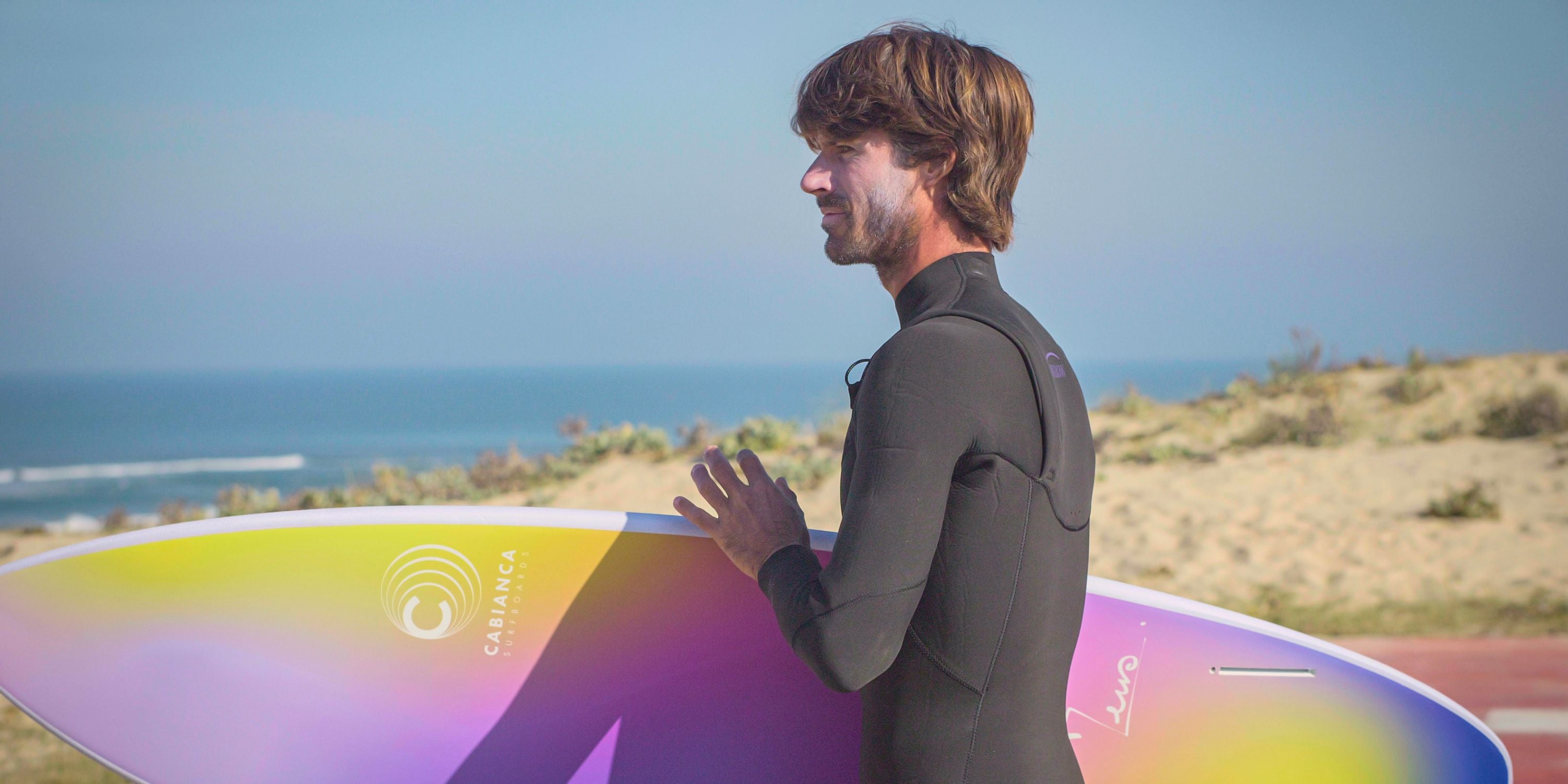 Un surfeur au visage souriant et avec de la crème solaire il porte une combinaison de surf oxbow noire et il est  en train de porter une de surf planche de surf shortboard multicolore et regarder le large sur la côte landaise